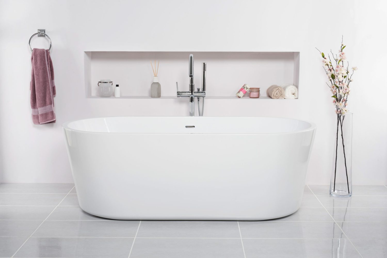 Bathtub in a white bathroom with flowers in a vase next to the tub and skincare products in the rack A bathtub in a white bathroom with flowers in a vase next to the tub and skincare products in the rack